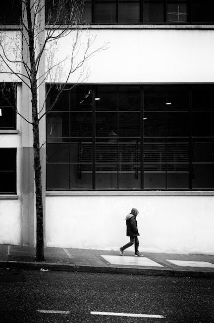 Person walks past a stark white building and a bare tree on a sidewalk. Black and white.