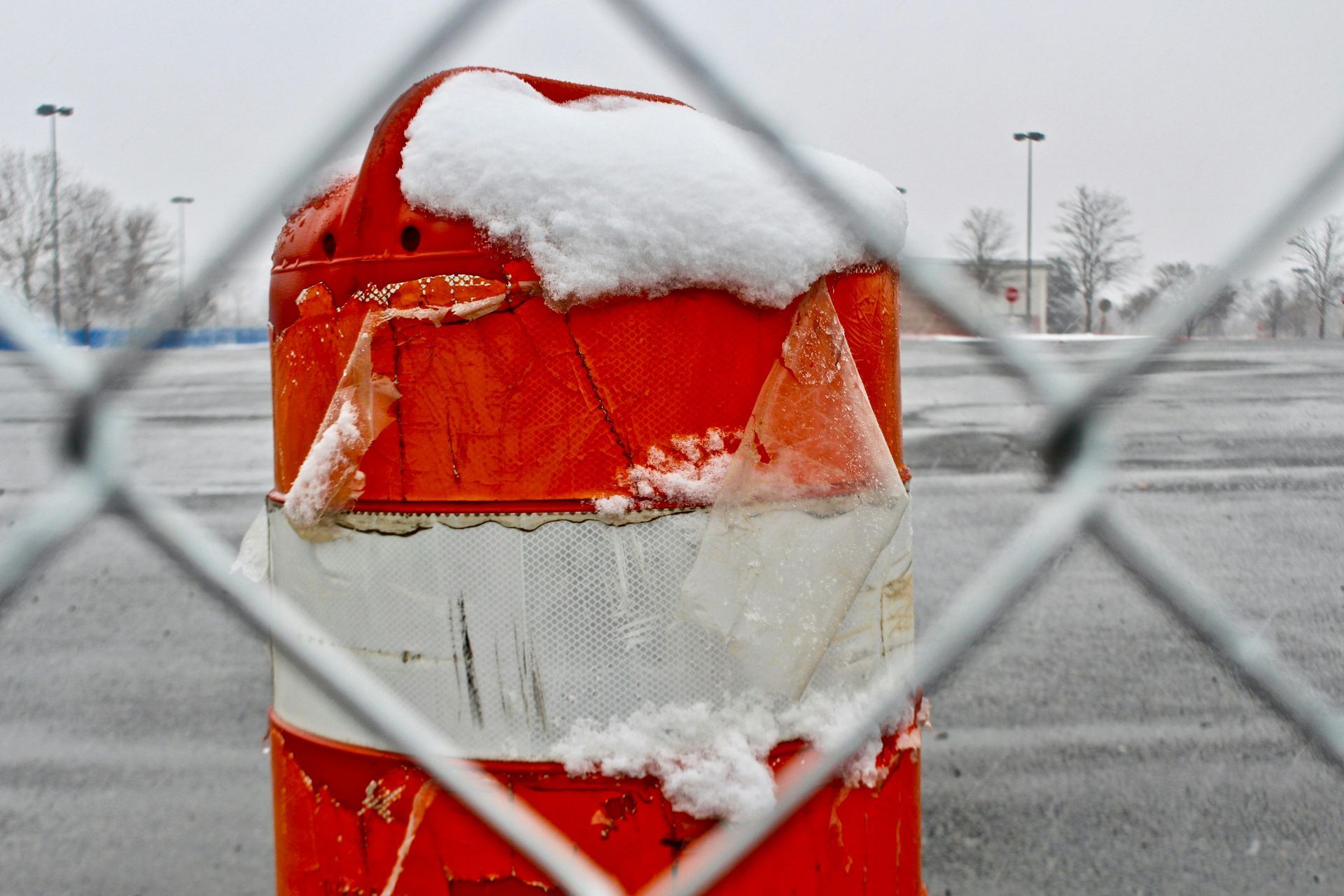 Orange traffic barrel with snow on top, seen through a chain-link fence in a parking lot.
