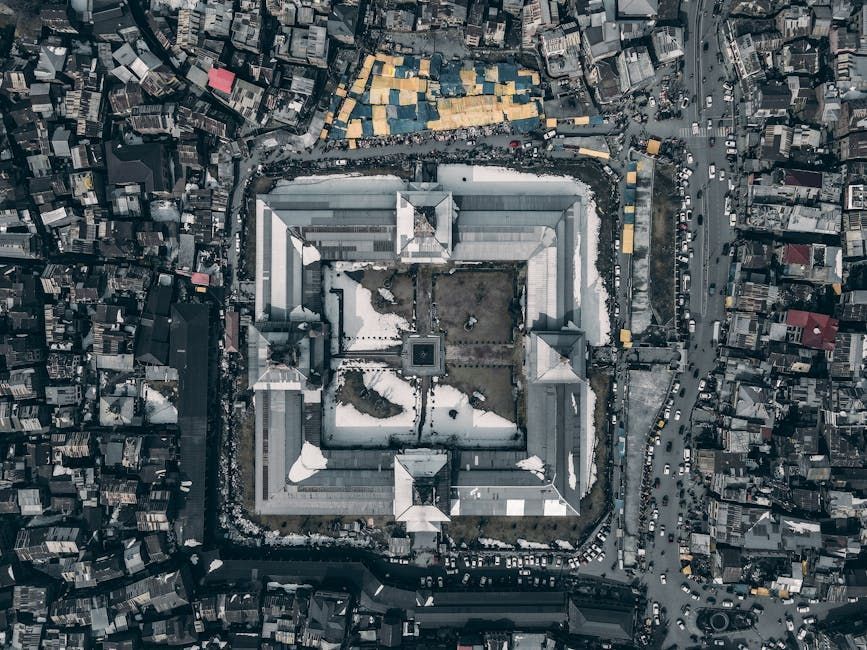 Overhead view of a large, square, white-roofed building surrounded by dense city structures and roads.