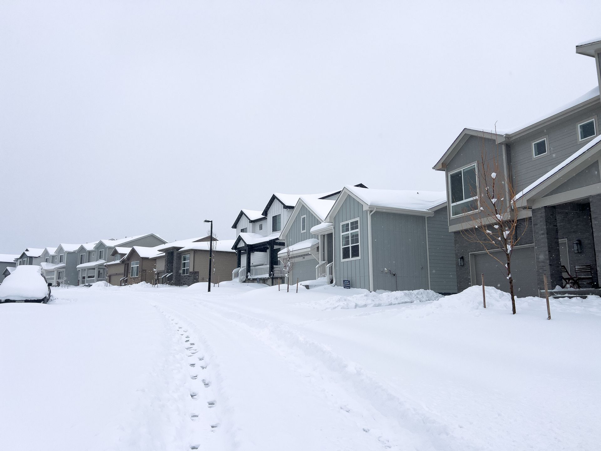 Snowy residential street with houses covered in snow. Footprints and a car visible.