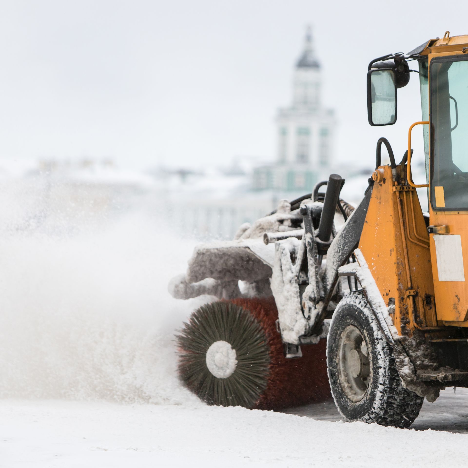Yellow snow sweeper clearing snow from a road, white winter setting.
