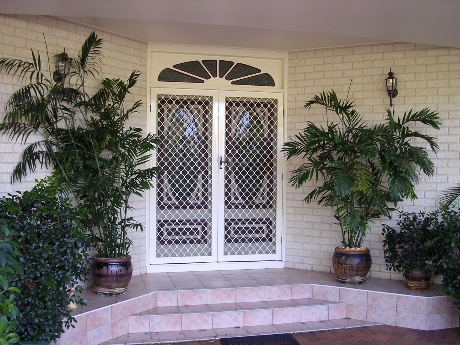 Front porch with white double doors, potted palms, and tiled steps beneath an arched window. — Advantage Screen And Blinds Rockhampton in Rockhampton, QLD