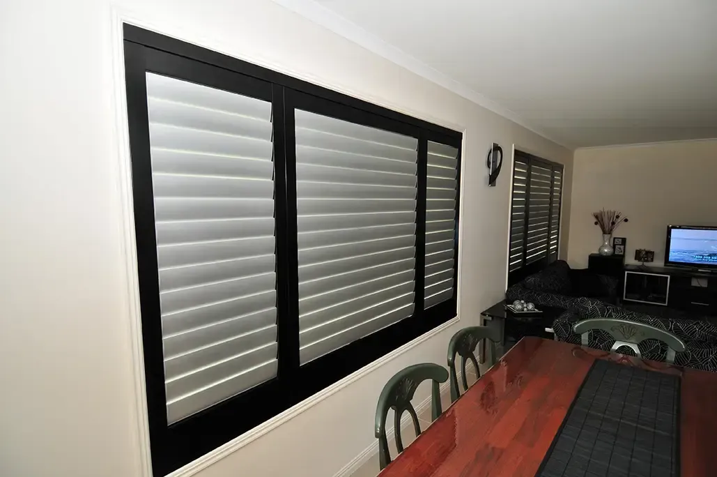 A Dining Room With a Large Window Covered by Horizontal Shutters, With a Wooden Table and Chairs Visible — Advantage Screen And Blinds Rockhampton in Rockhampton, QLD