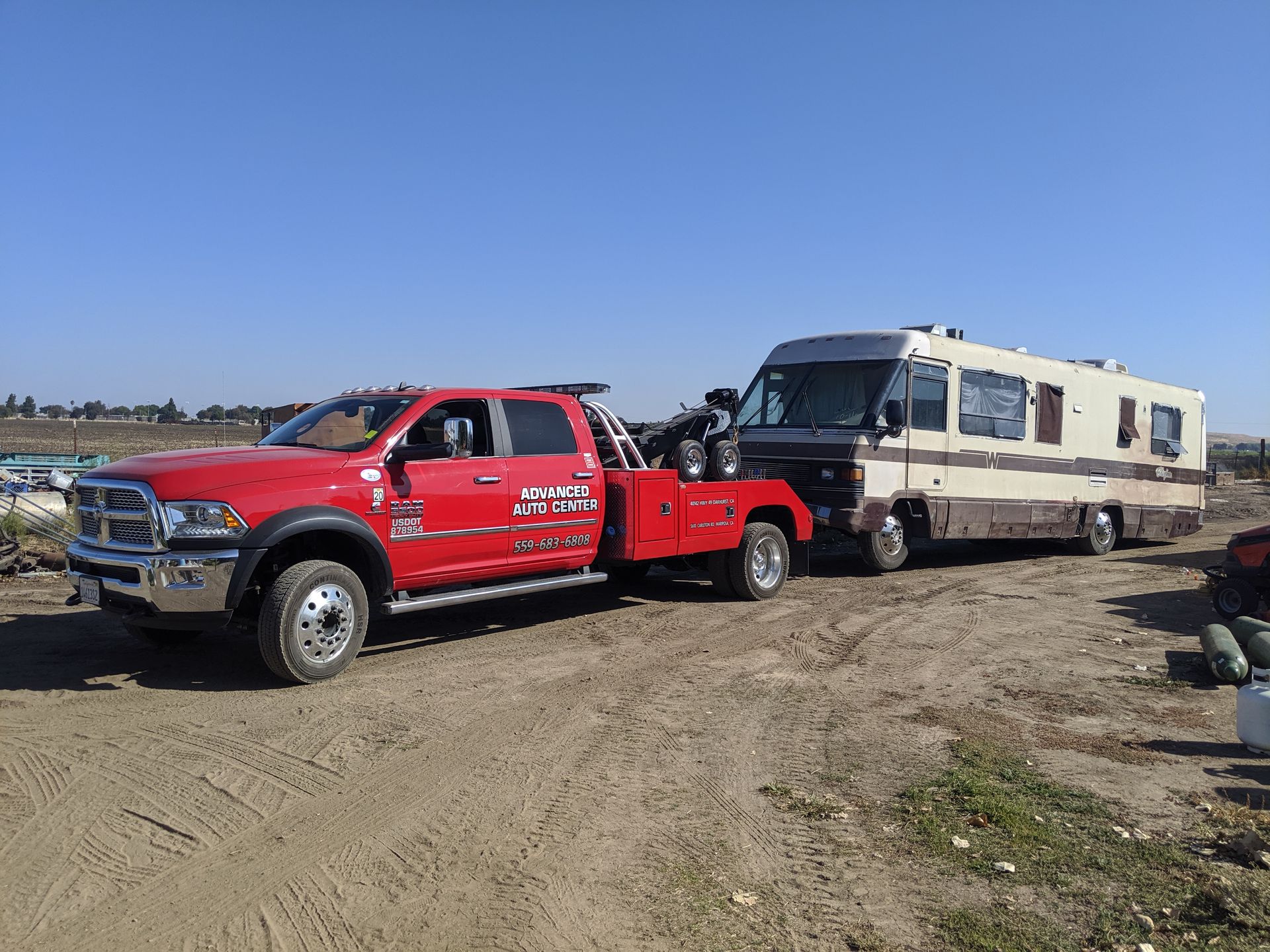 Red tow truck towing a beige and brown RV on a dirt lot under a blue sky.