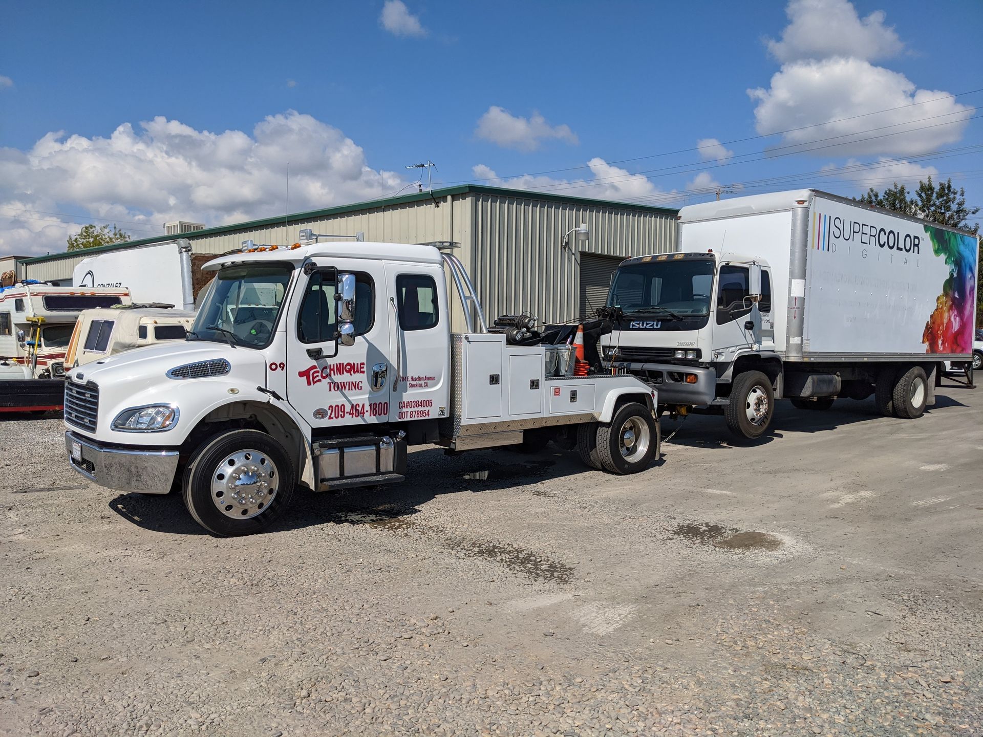 White tow truck towing a box truck on a gravel lot under a blue sky.