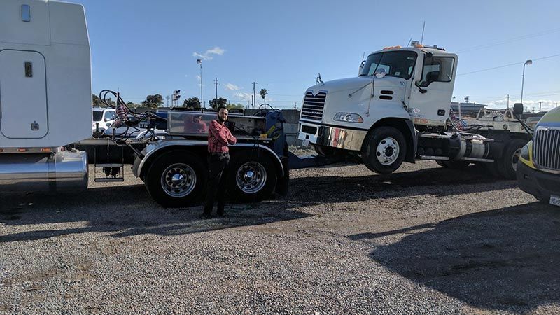 A man is standing next to a semi truck in a gravel lot.