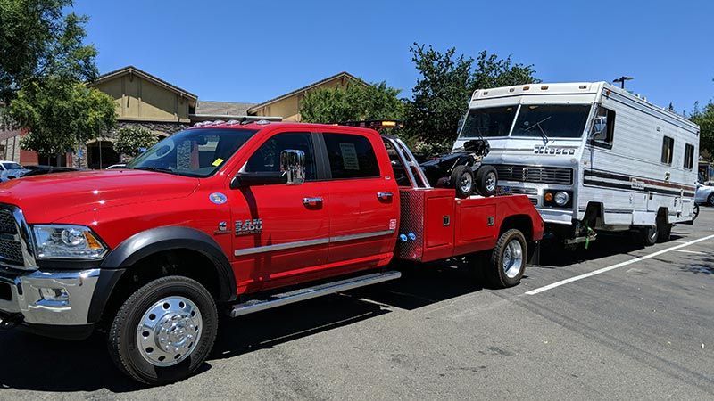 A red tow truck is towing a white rv down a street.