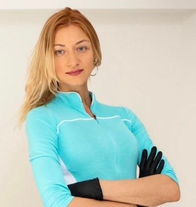 A person with blonde hair stands with arms crossed, wearing a turquoise long-sleeved shirt and black gloves.