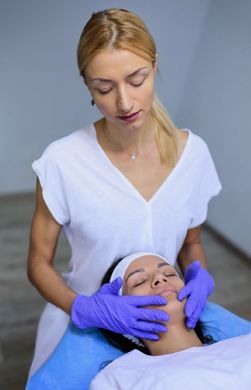 A practitioner wearing blue gloves performs a facial massage on a client lying down in a treatment room.