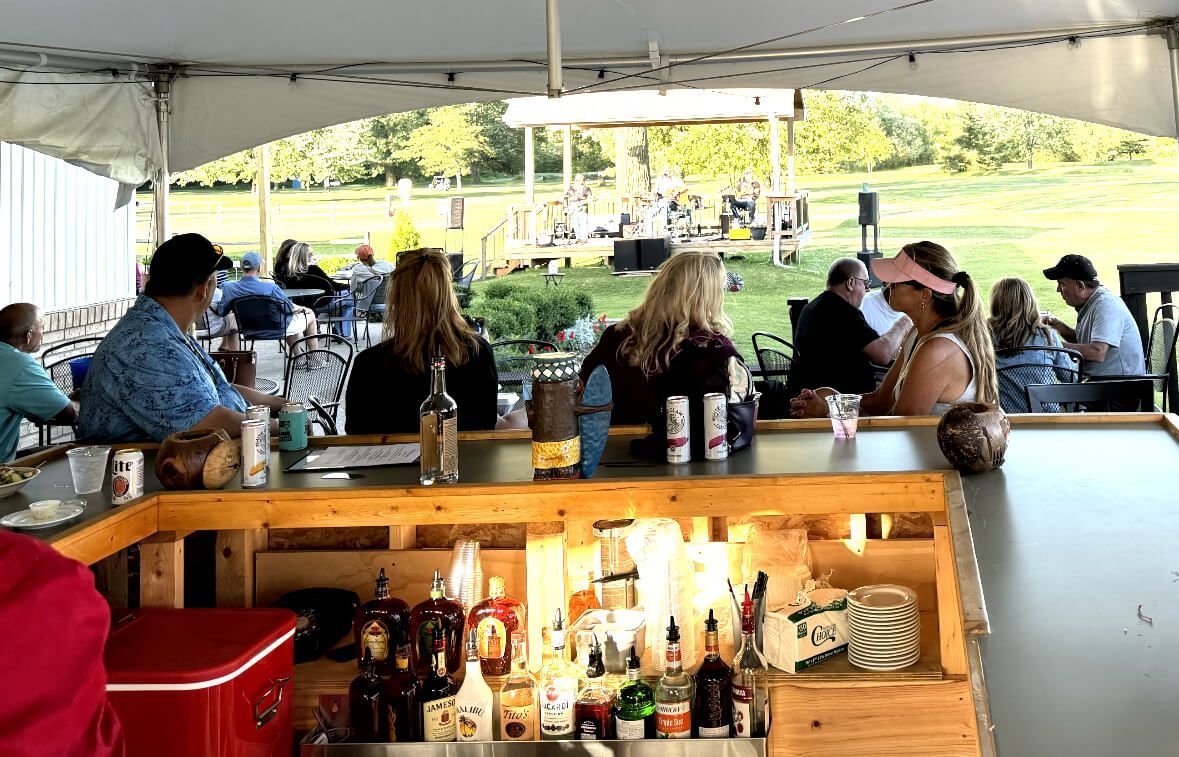 A group of people are sitting at a bar under a tent at Little Camille's by the Green restaurant in Casco, Michigan.