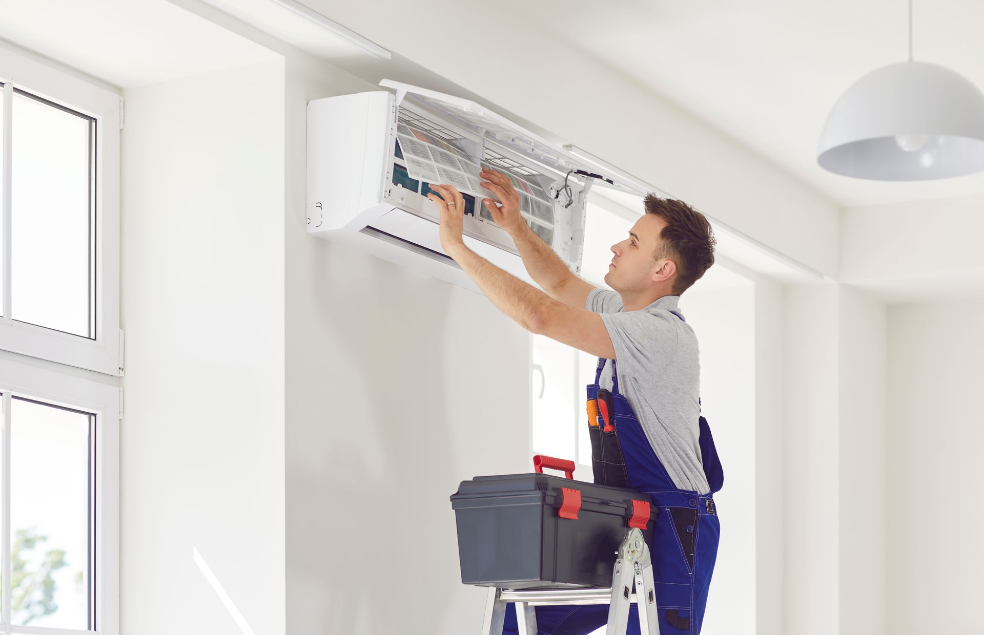 Man in blue overalls servicing a white air conditioner, holding filters, with a toolbox on a stepladder.