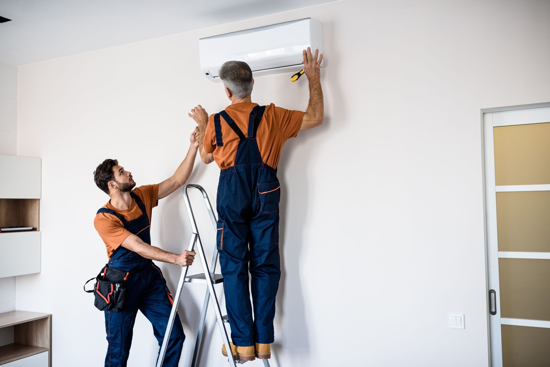 Two workers installing an air conditioner on a wall using a ladder.