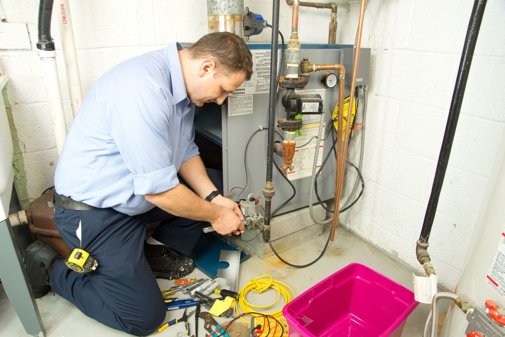Plumber kneeling, working on a furnace in a basement. Tools and pink bin are on the floor.