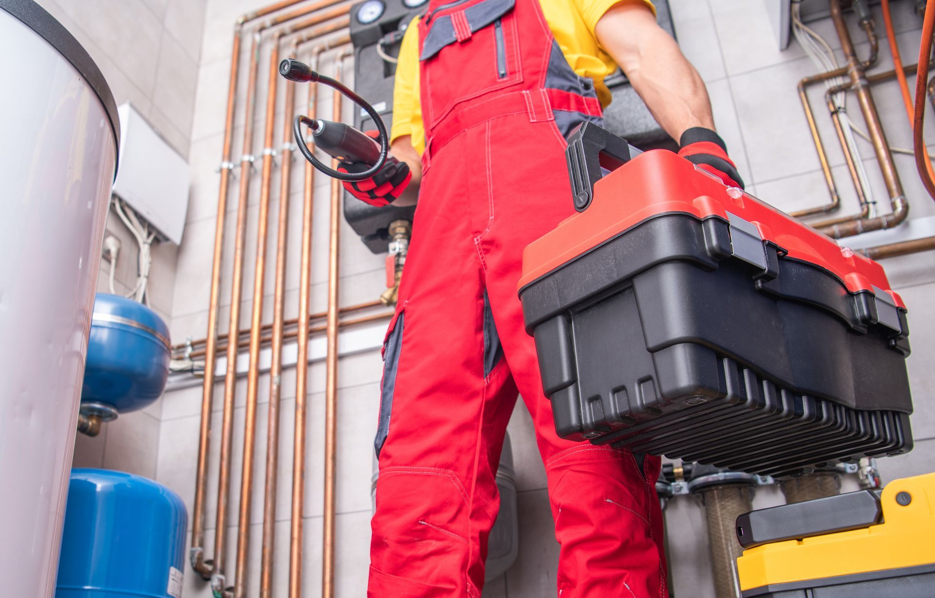 Plumber in red overalls holding a toolbox and instrument, standing in a mechanical room with copper pipes.