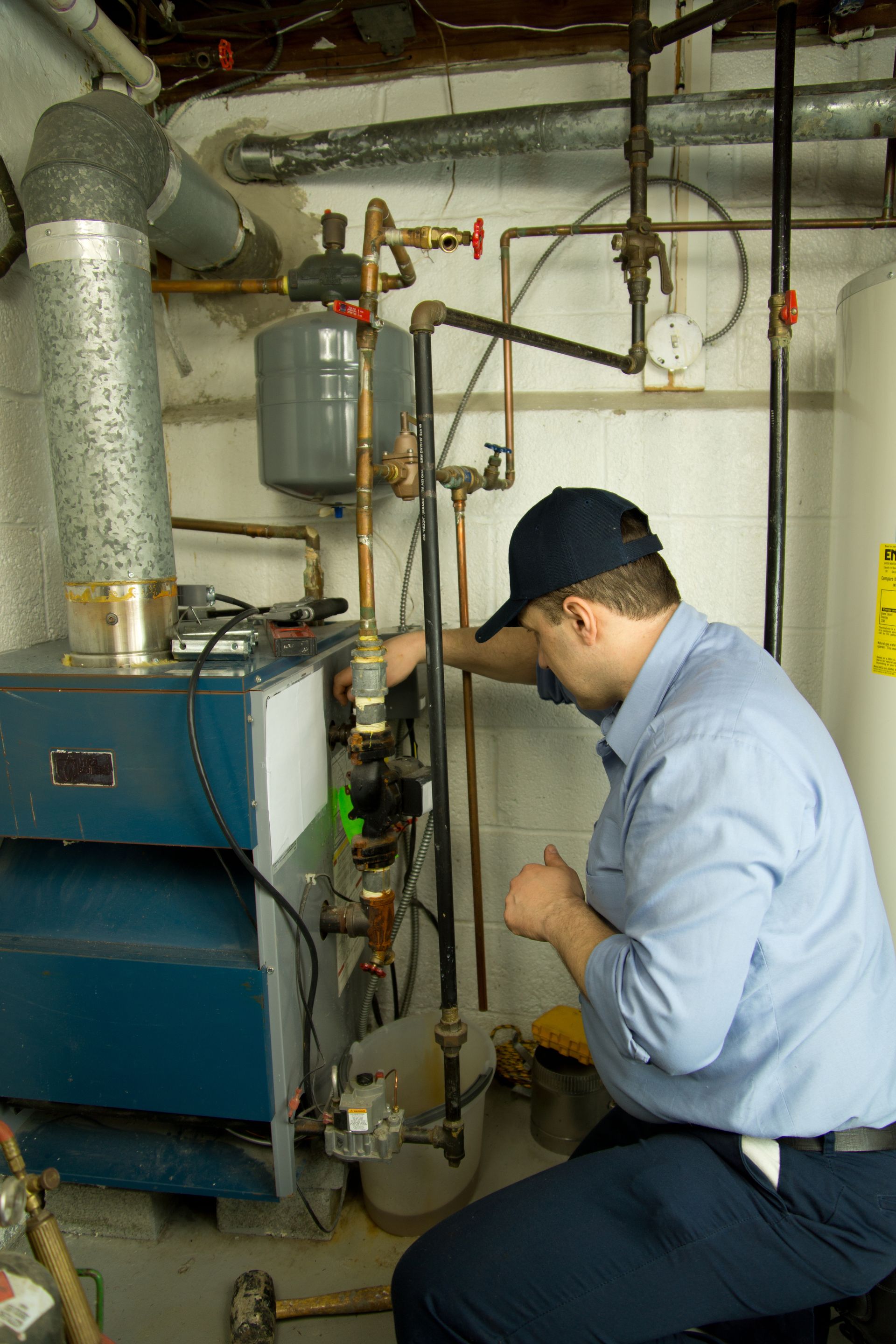 Plumber in a blue shirt and cap working on a furnace with various pipes and gauges.