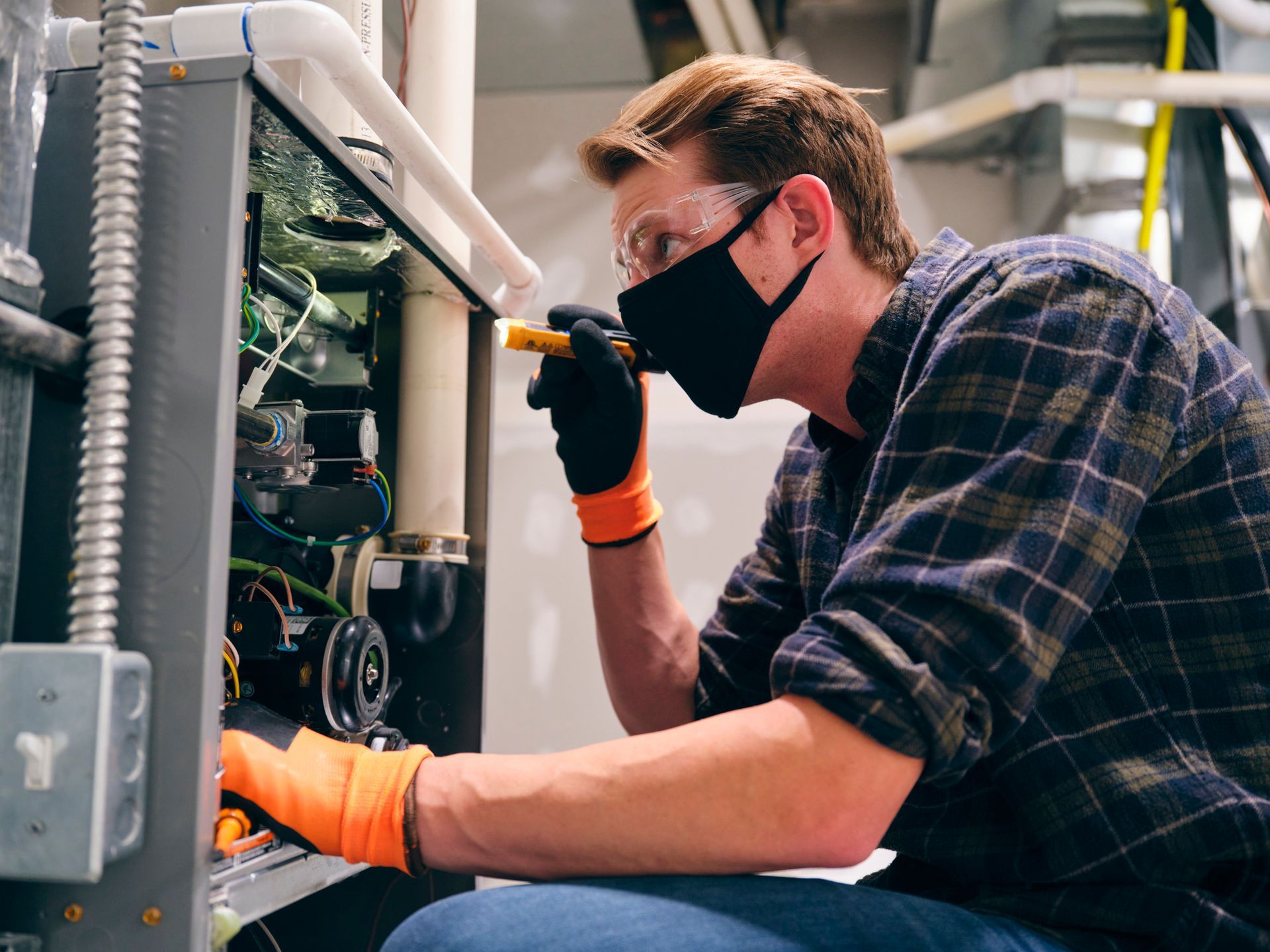 HVAC technician in a face mask and safety glasses, inspecting equipment with a flashlight.