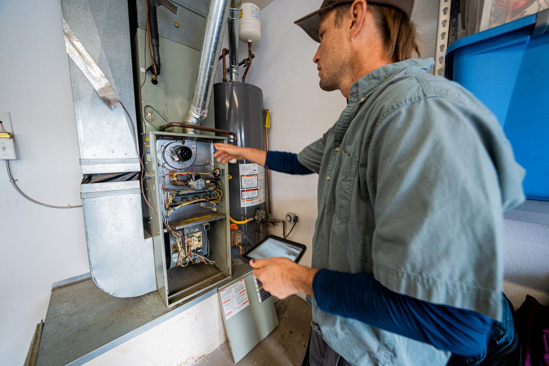 HVAC technician inspecting furnace, holding tablet. Indoor setting with water heater, blue bin.