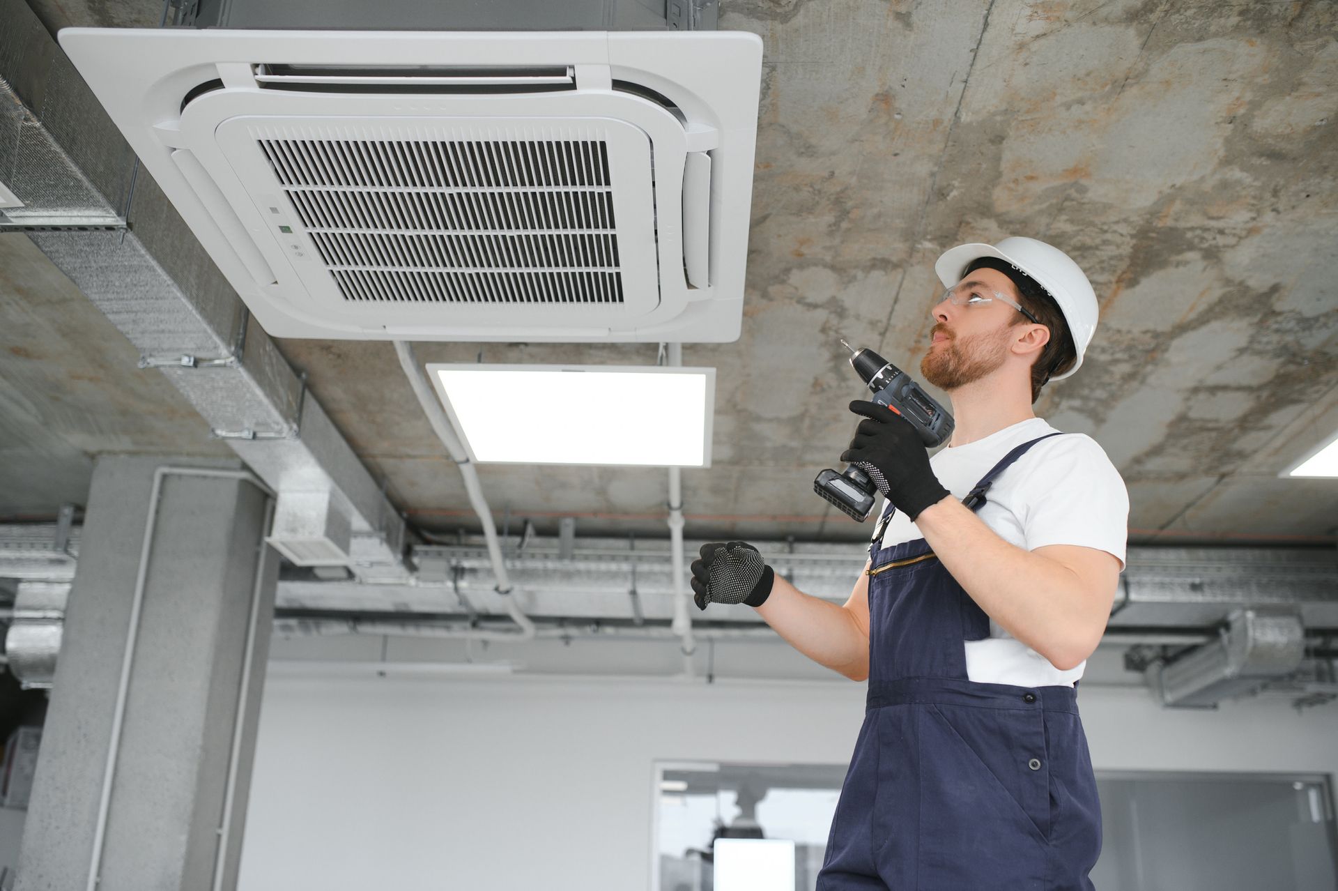 HVAC technician in overalls, hard hat, using a drill to install ceiling-mounted air conditioning unit.