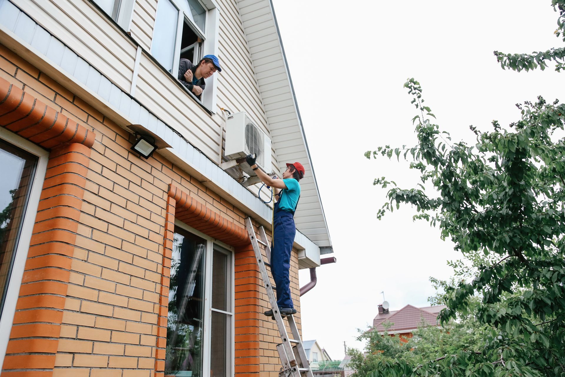 Two workers installing an AC unit outside a two-story brick house, one on a ladder, the other in a window.