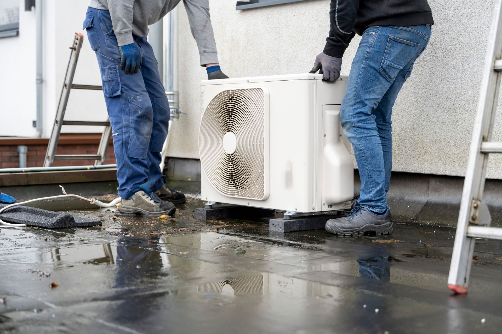 Two workers lifting an air conditioning unit on a wet rooftop.