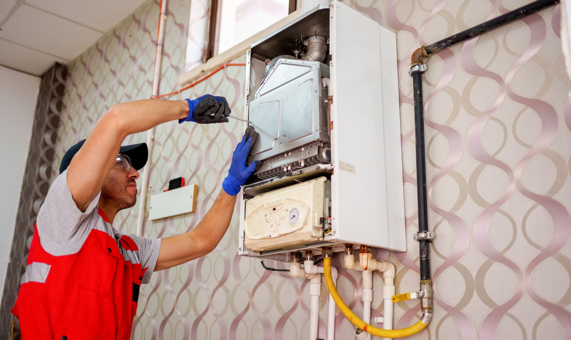 Technician in blue gloves repairs a wall-mounted boiler, wearing a red vest.