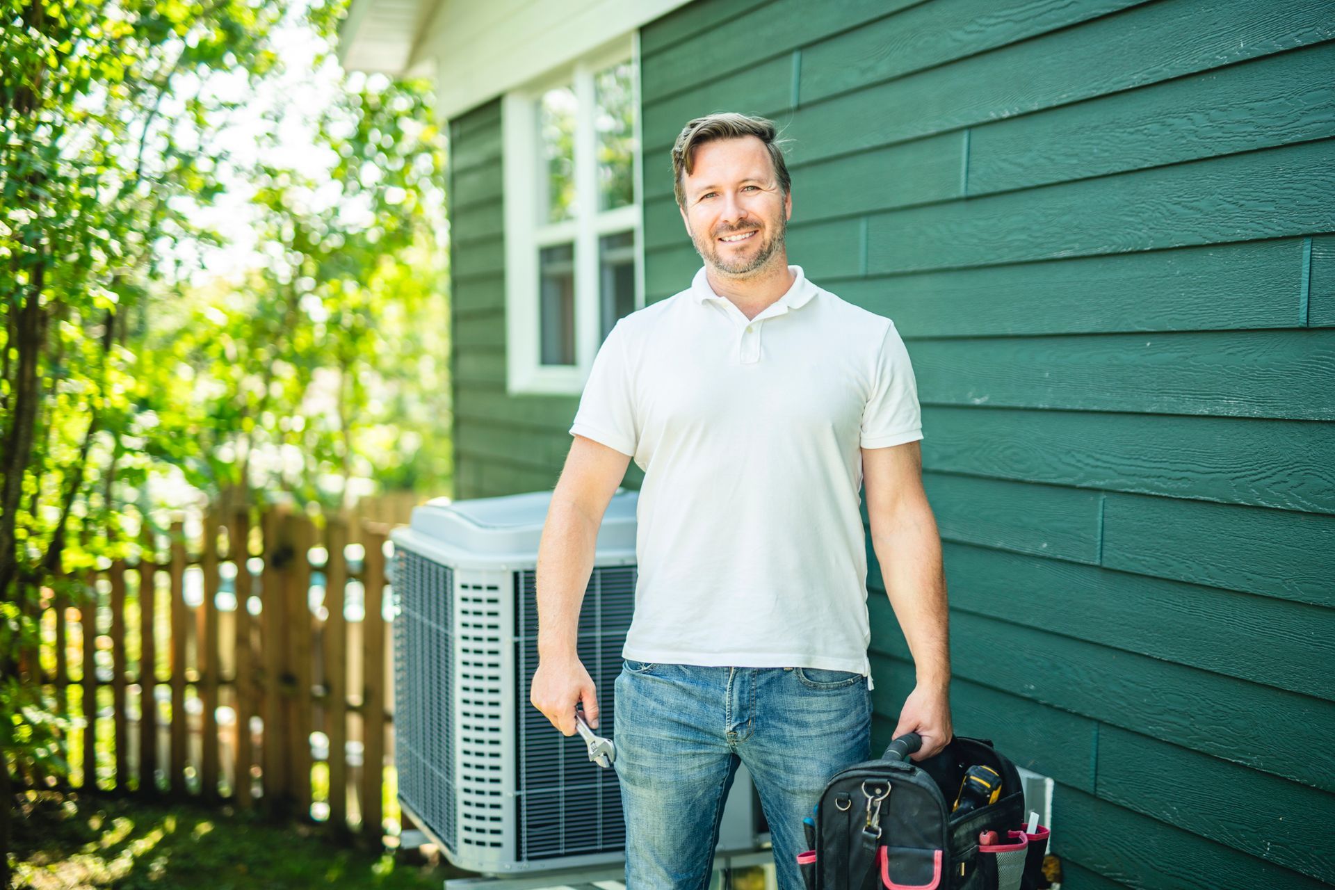 Smiling man with tools stands by an air conditioner near a green house.