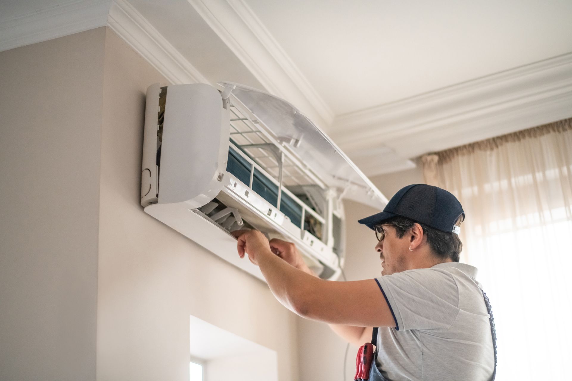 Man in cap repairs air conditioner on a wall.