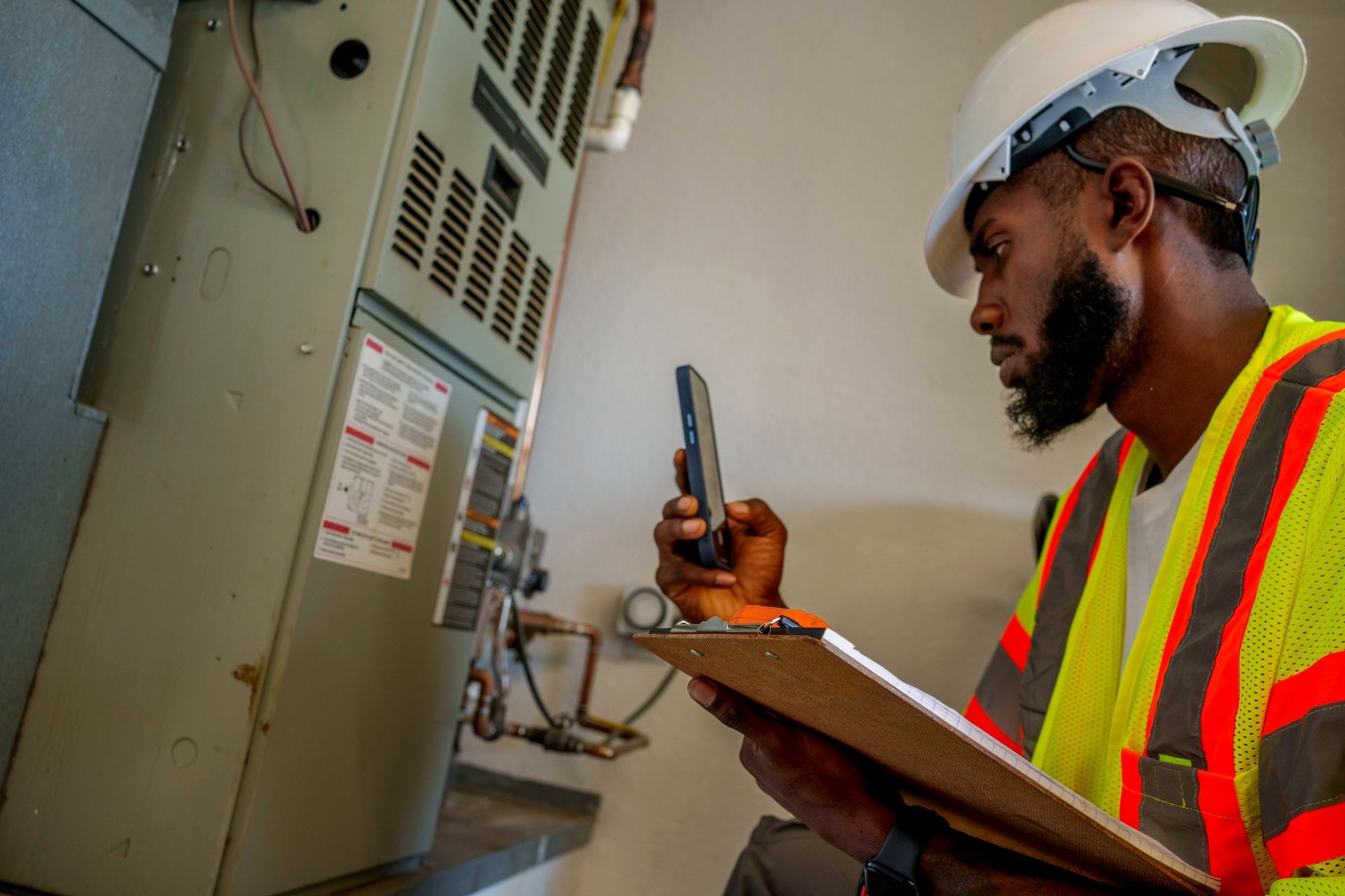 HVAC technician inspecting unit, wearing a hard hat and safety vest, using a phone, and holding a clipboard.