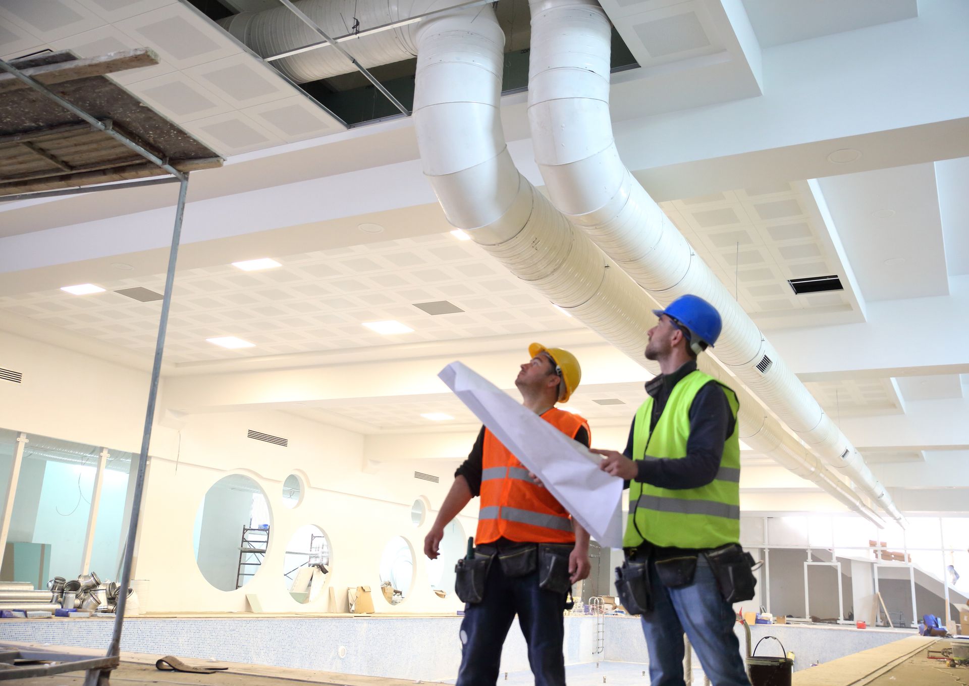 Two construction workers in safety vests inspect HVAC ducts in a building under construction, one holding blueprints.