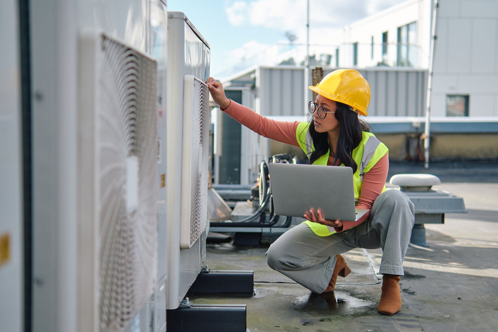 Person in a hard hat and safety vest inspecting HVAC equipment on a rooftop, using a laptop.
