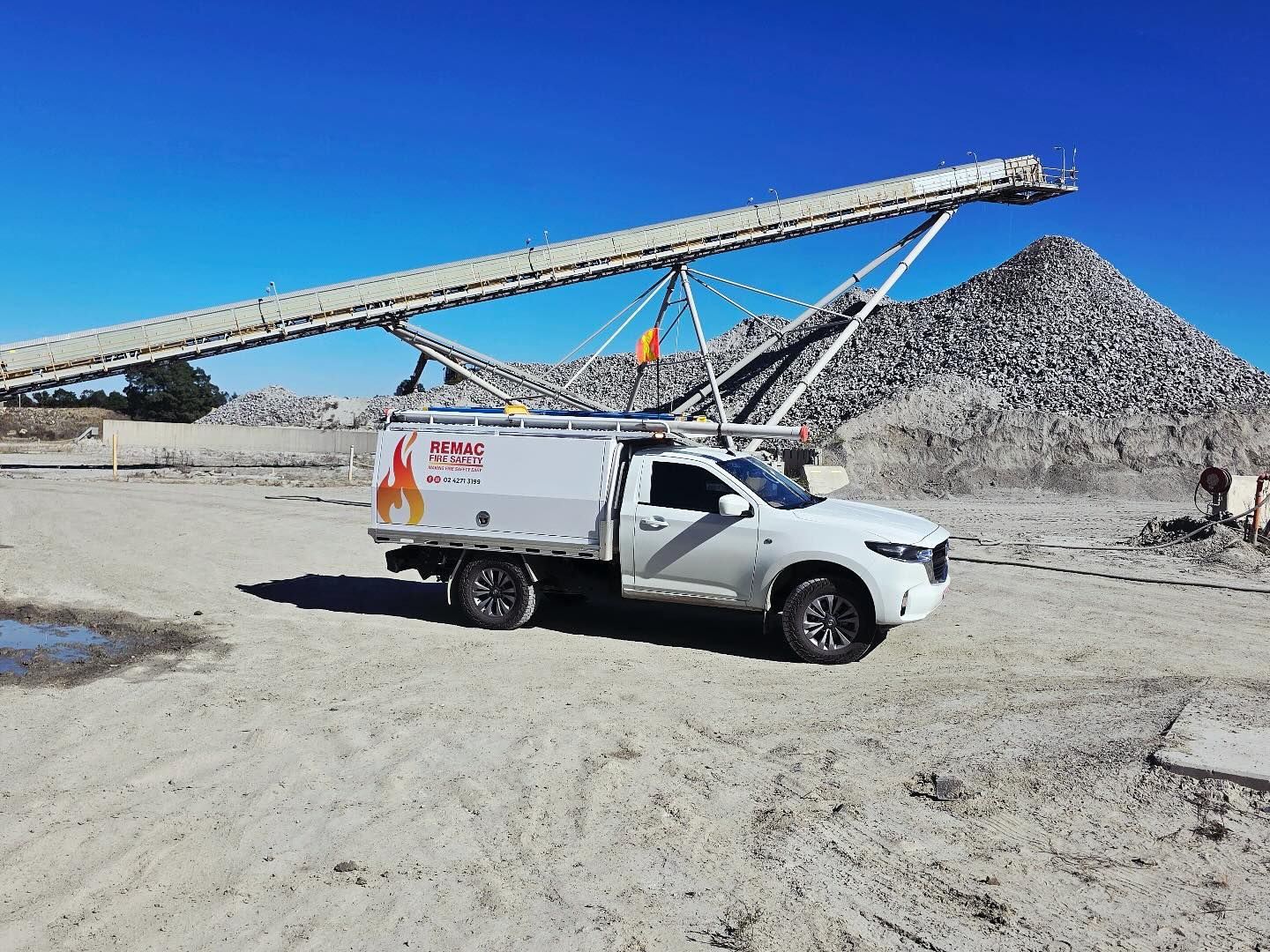 A white truck is parked in the dirt in front of a conveyor belt.