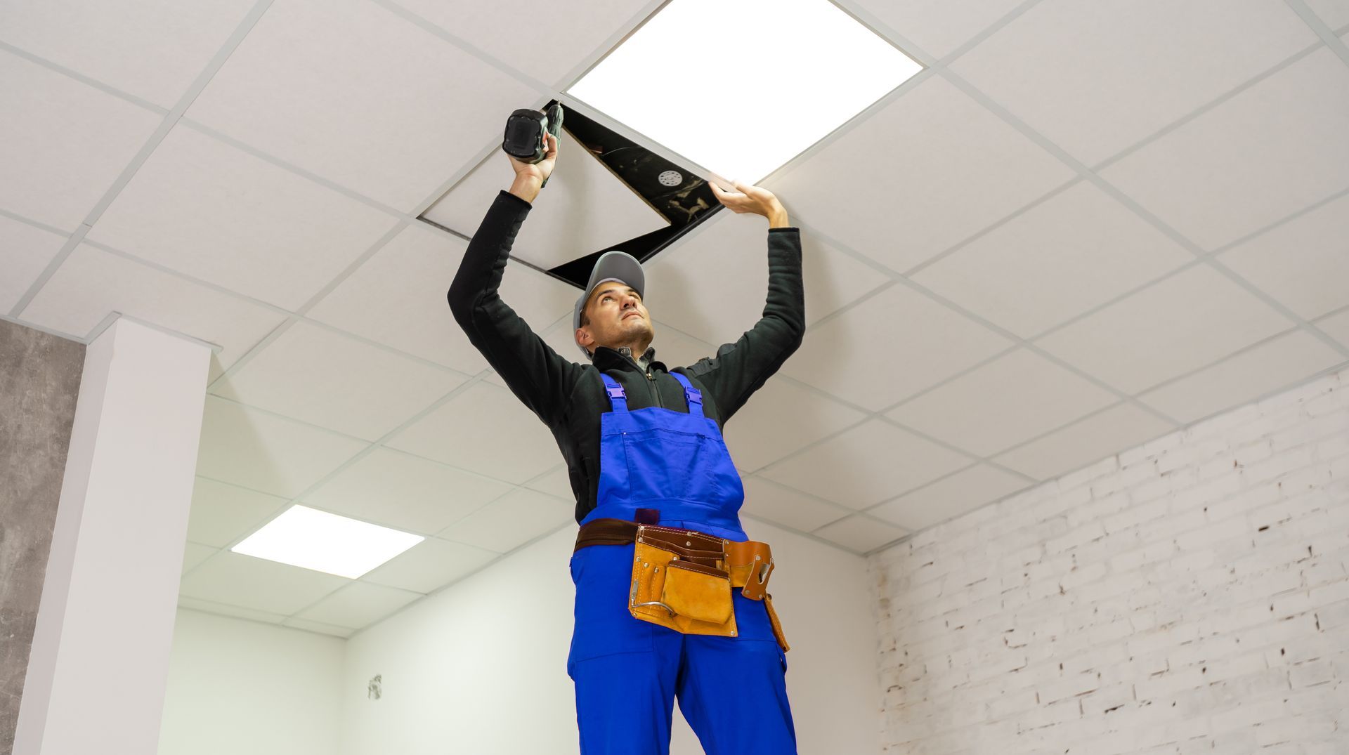 Worker installing a panel in a drop ceiling using a drill.