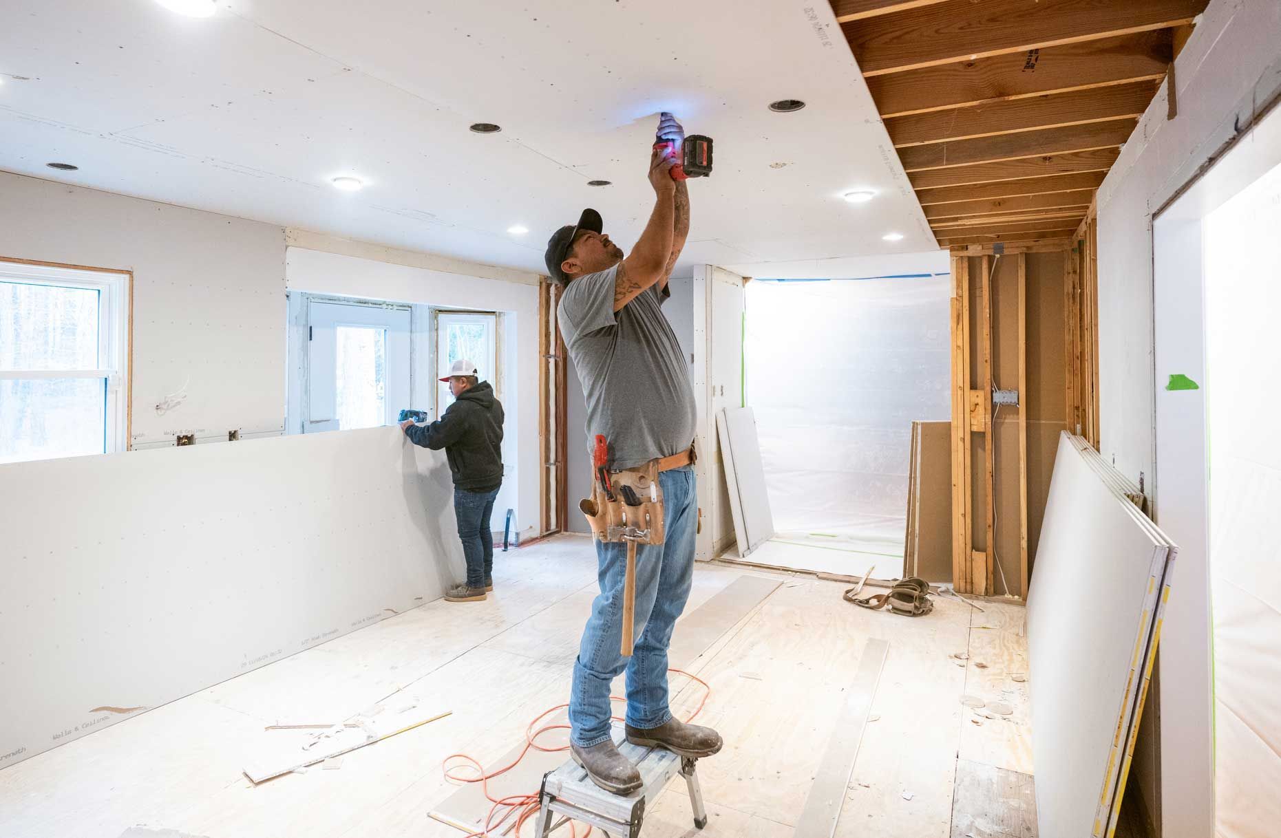 Two workers install drop ceiling panels in room under renovation with tools and recessed lights.