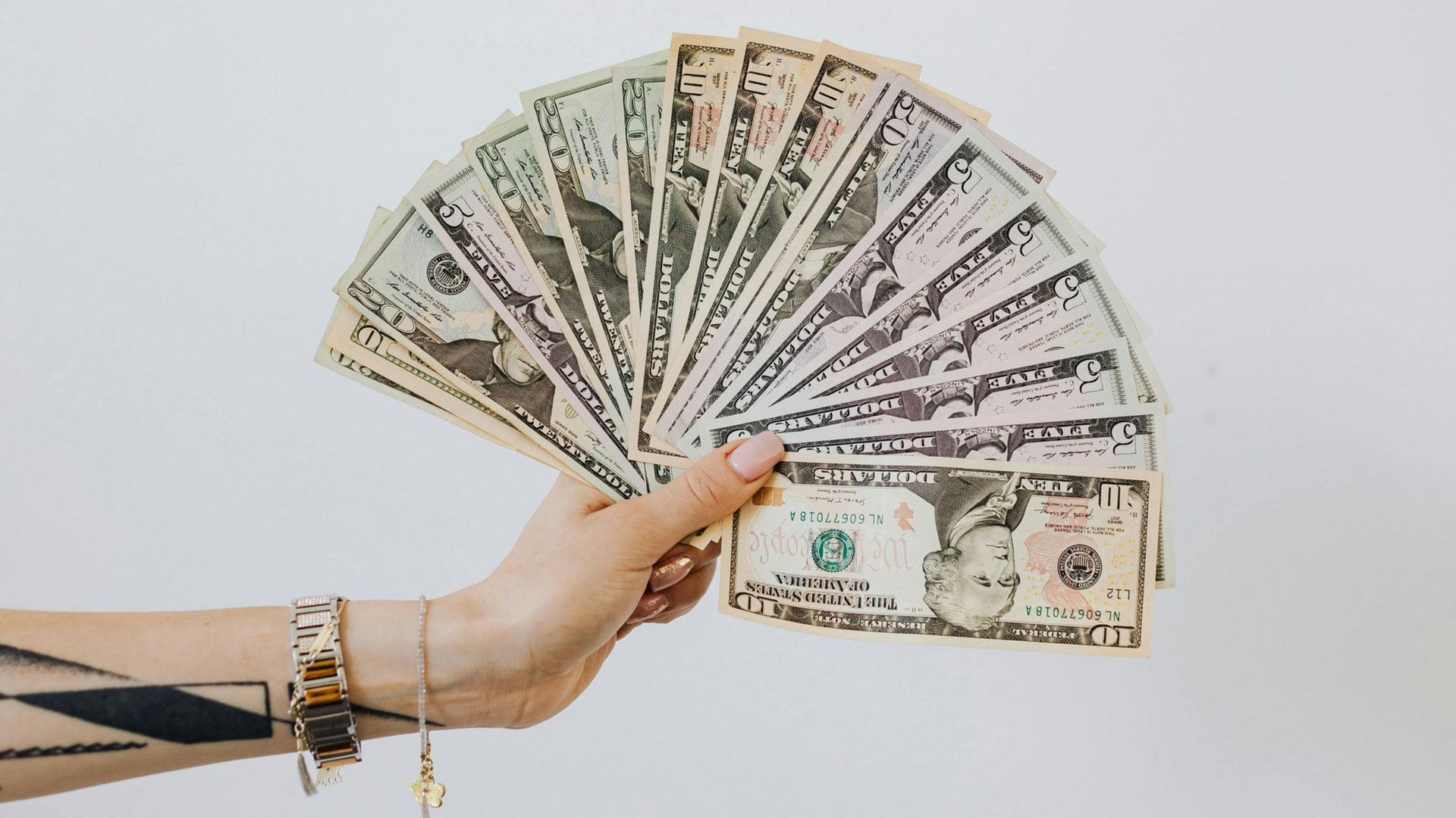 Hand holding a fan of US dollar bills against a white background, with a tattoo on the arm.