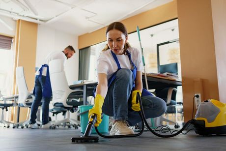 A woman is cleaning the floor of an office with a vacuum cleaner.