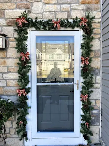 A front door decorated for Christmas with a green garland featuring red plaid bows and pinecones against a stone wall.