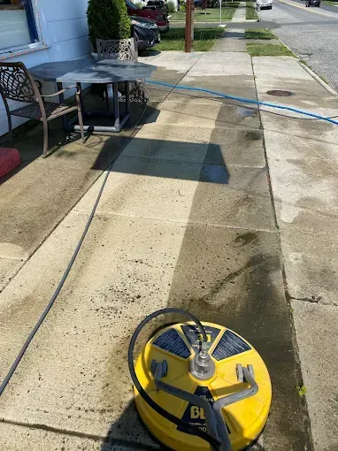 A yellow surface cleaner sits on a concrete sidewalk, showing a clean, light-colored path next to darker, uncleaned pavement.