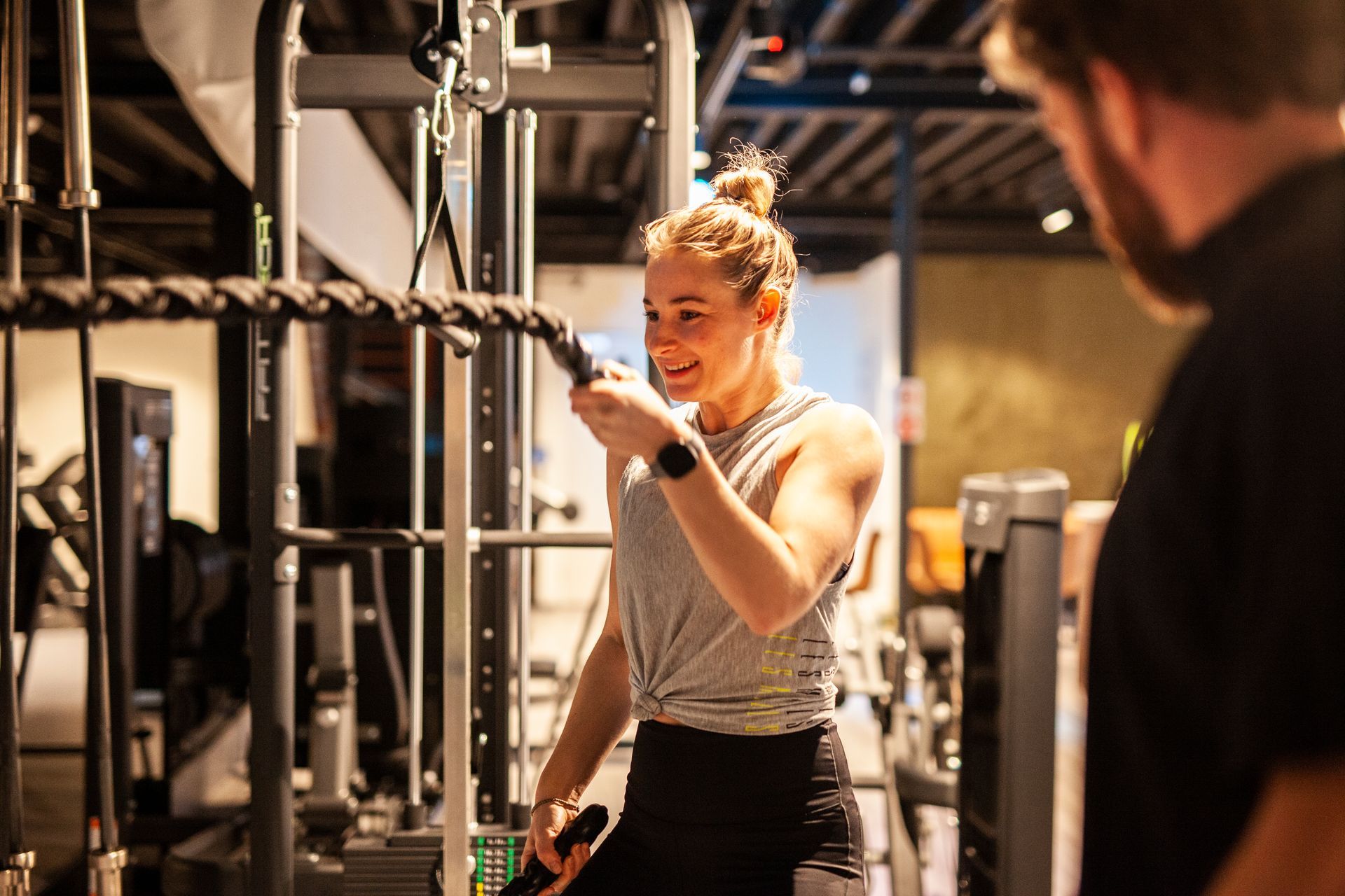 Een vrouw tilt een halter op in de sportschool, terwijl een man toekijkt.