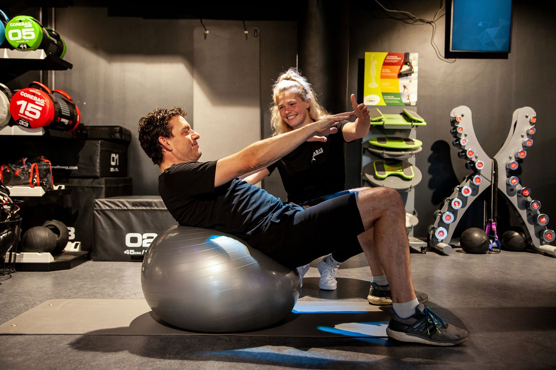 Een man doet sit-ups op een oefenbal in een sportschool, terwijl een vrouw toekijkt.