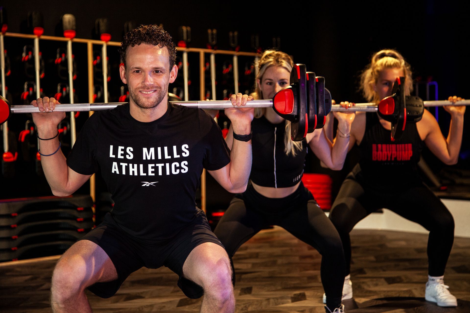 Een man en twee vrouwen hurken met halters in een sportschool.