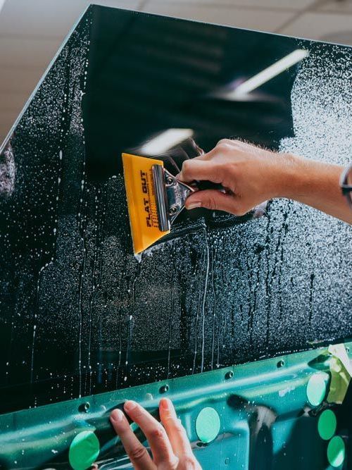 Person applying tint film to a car window with a squeegee. Green window and vehicle trim visible.