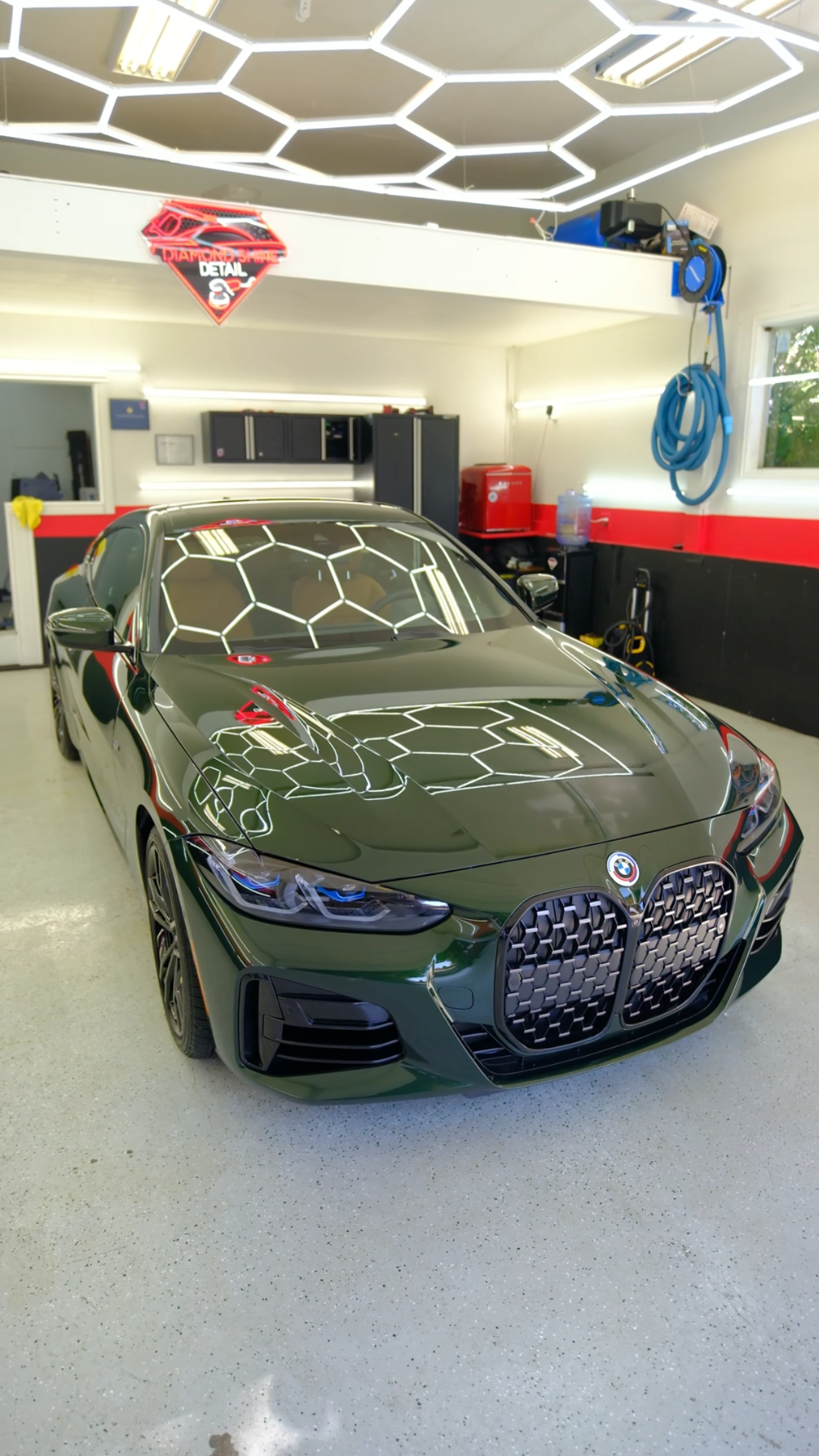 Green BMW car in a garage with honeycomb ceiling lights, black trim, and tools.