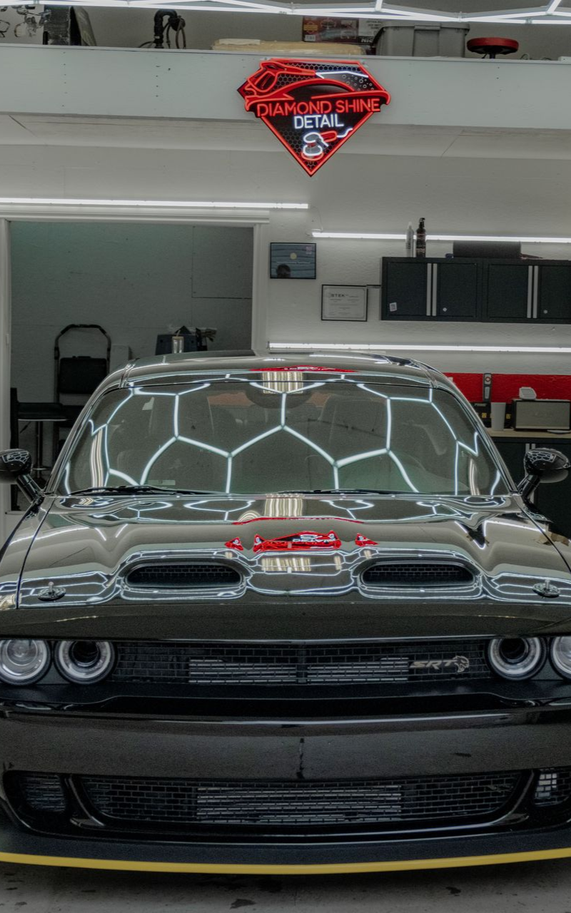 Dark Dodge Challenger car, illuminated in a detailing shop, with reflective lights on the windshield.