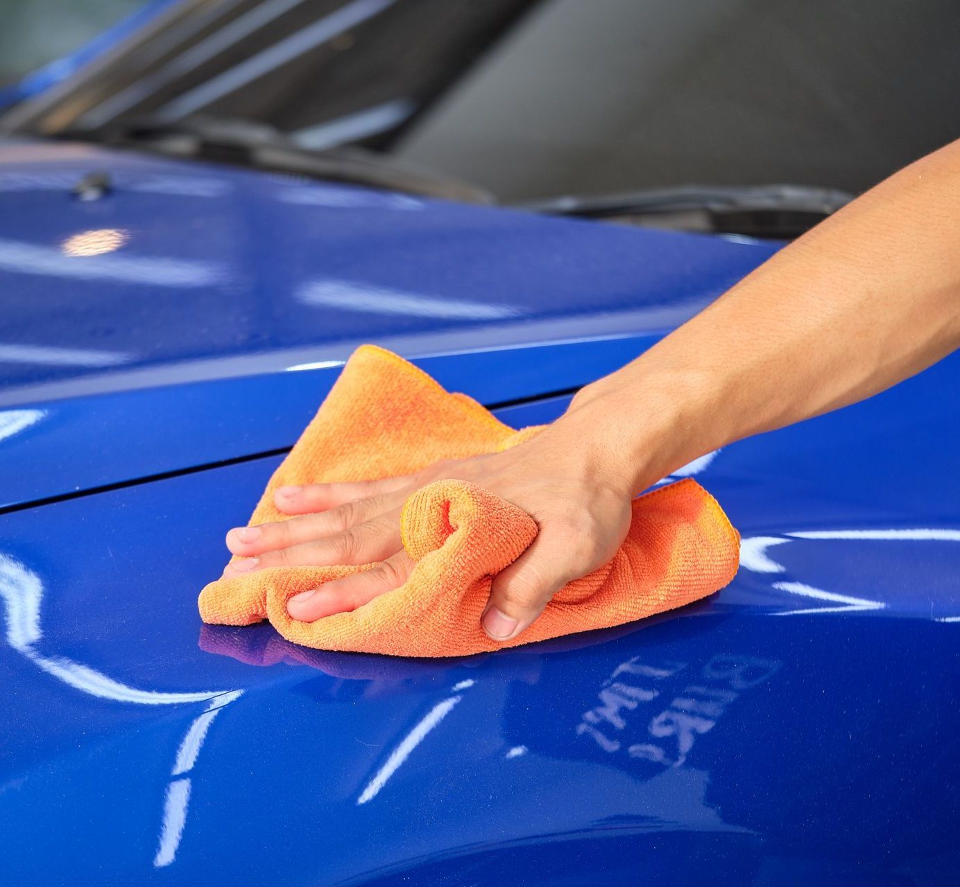 Hand wiping a bright blue car hood with an orange microfiber cloth.