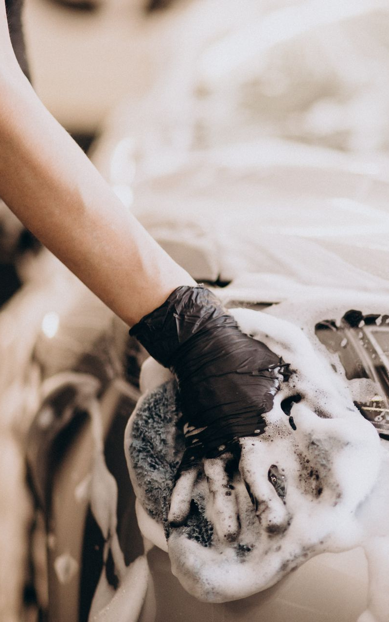 Hand wearing a black glove washing a car with foamy soap.
