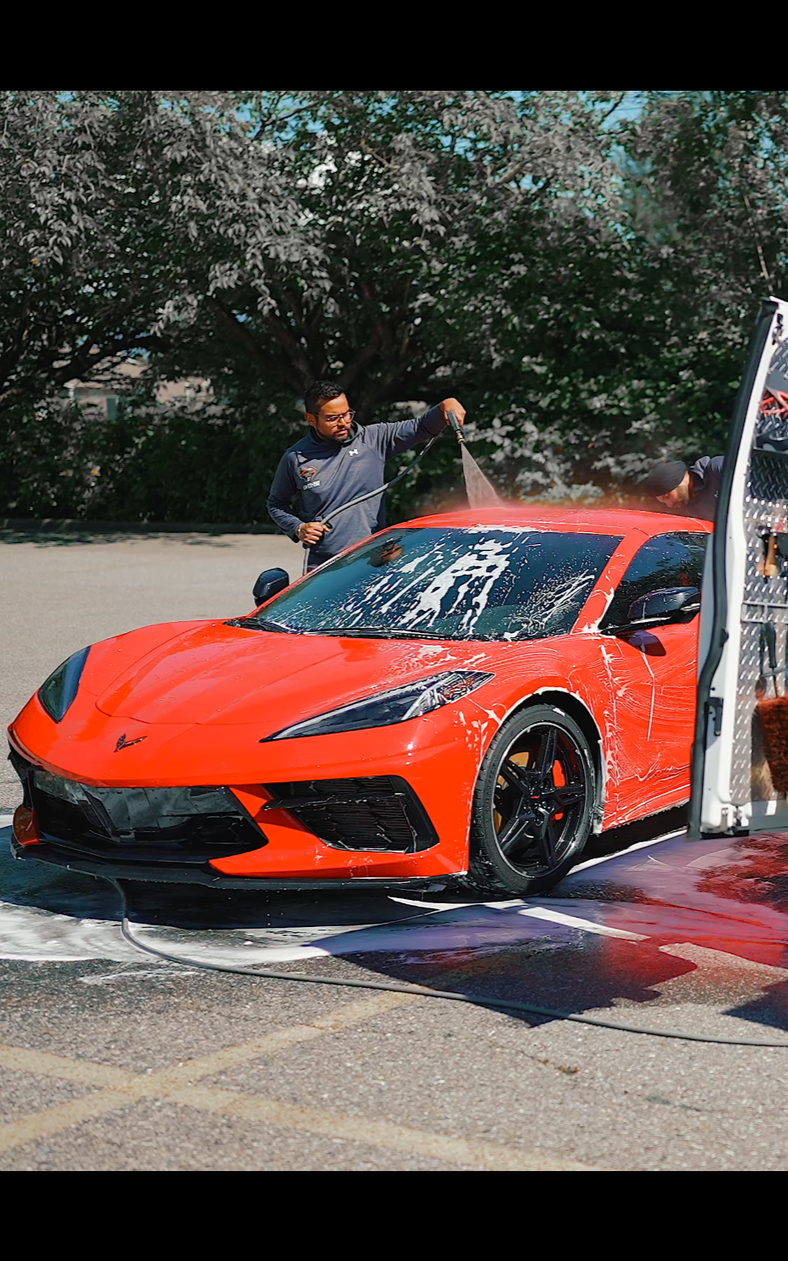 Man washing a bright red sports car with white foam, outdoors. Car door is open.