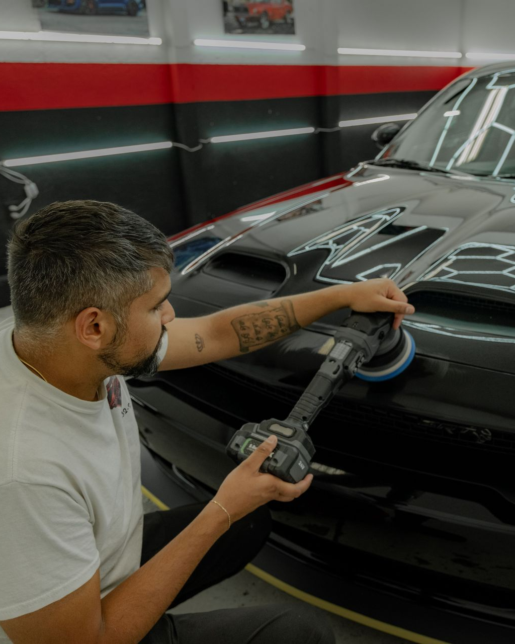 Man polishing a black car hood with a machine in a well-lit shop.