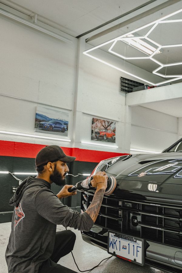 A person polishes a black car's hood with a machine in a well-lit garage.