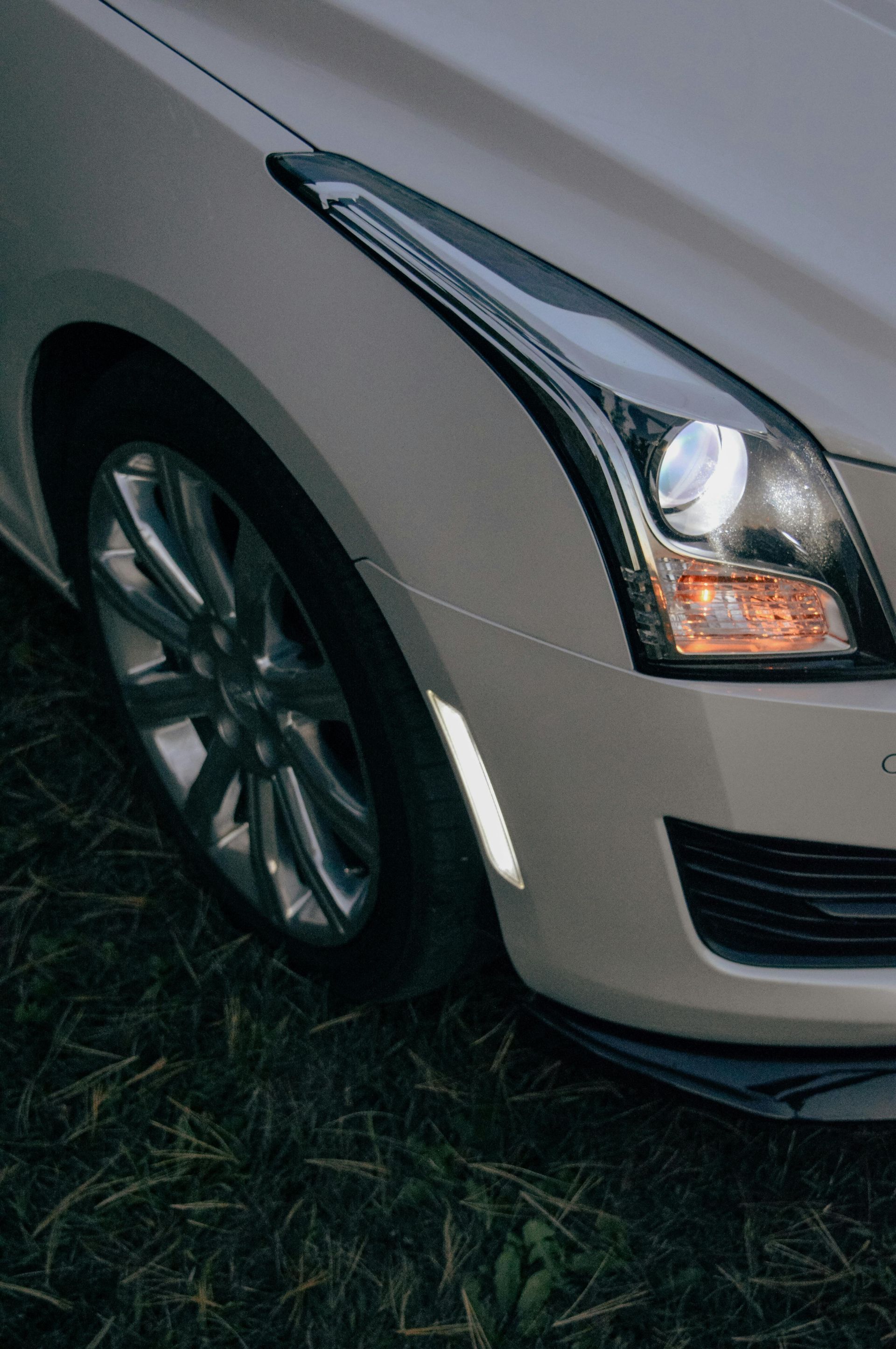 White Cadillac ATS front quarter, headlights illuminated, parked on grass.