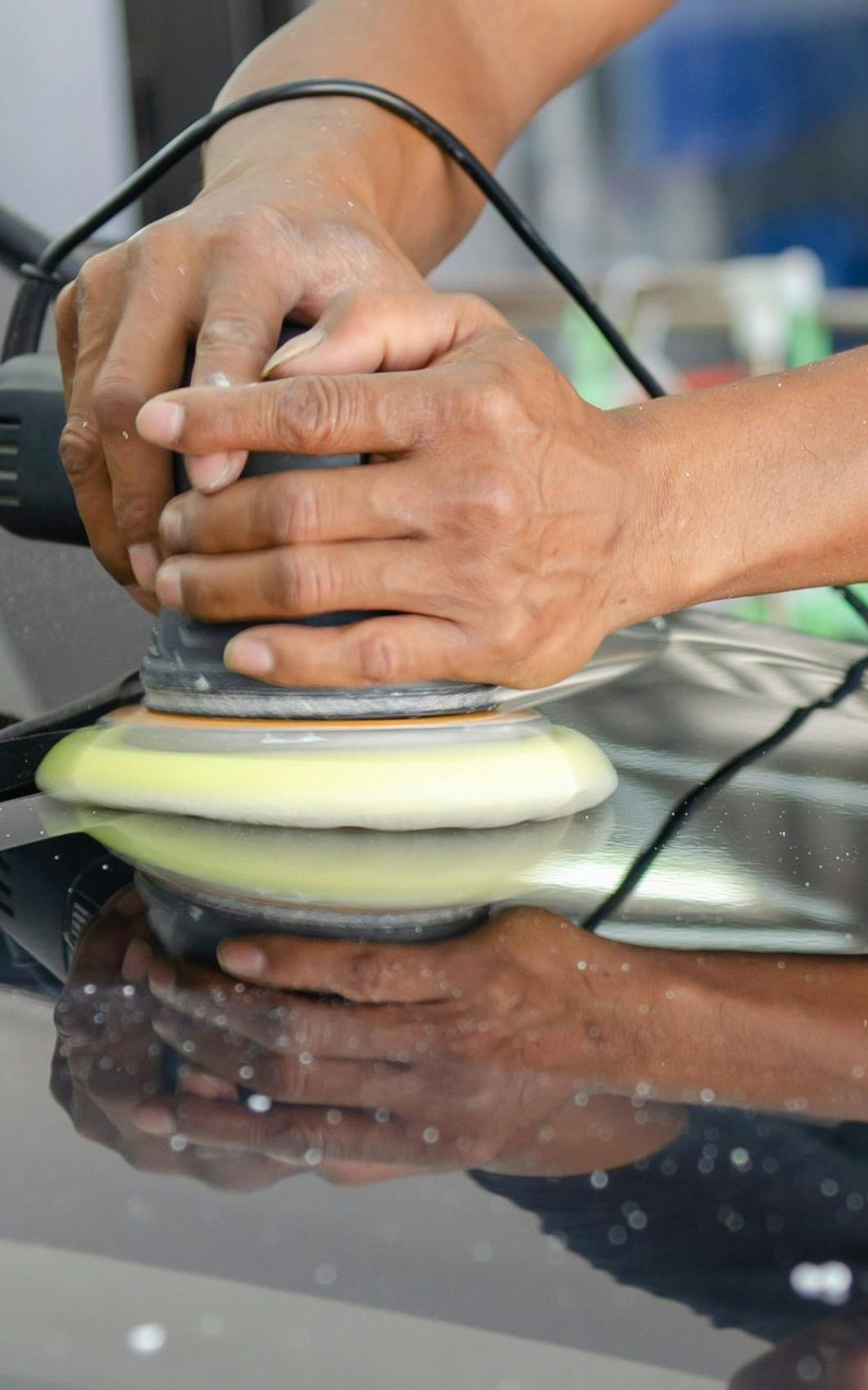 Hands using a buffer tool on a black car surface, reflecting the tool.